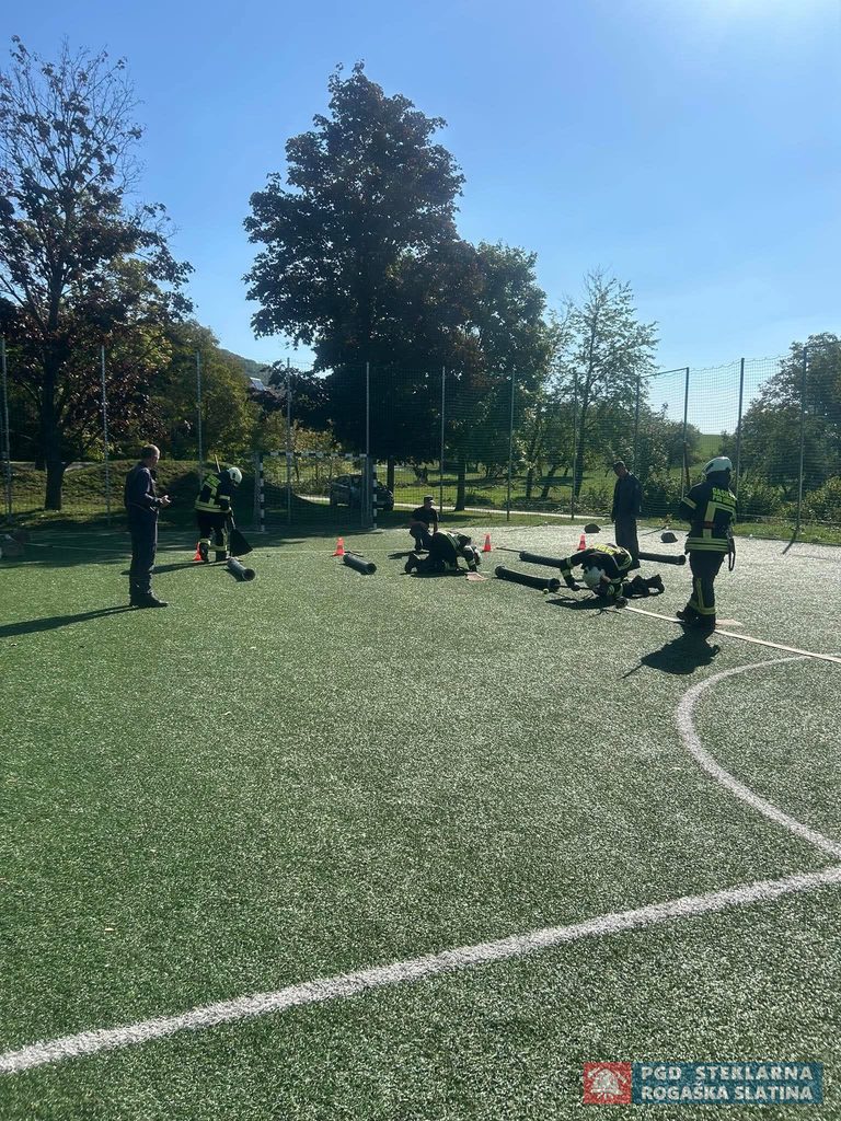 Gasilska ekipa pripravlja cevi in opremo na športnem igrišču.